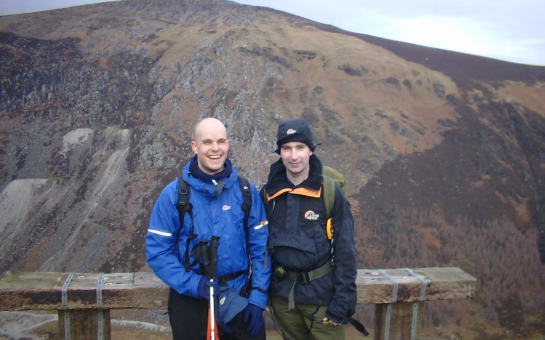 Mark Pollock and John ORegan stand before a mountainous landscape. Mark is wearing a blue jacket, holding two poles and John is wearing a navy jacket and hat. 