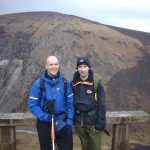 Mark Pollock and John ORegan stand before a mountainous landscape. Mark is wearing a blue jacket, holding two poles and John is wearing a navy jacket and hat. 