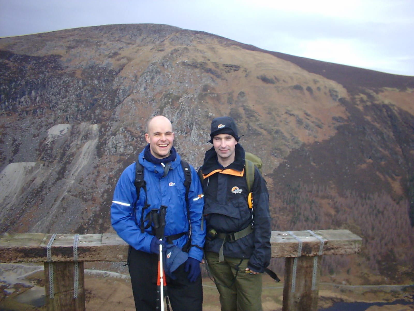 Mark Pollock and John ORegan stand before a mountainous landscape. Mark is wearing a blue jacket, holding two poles and John is wearing a navy jacket and hat. 