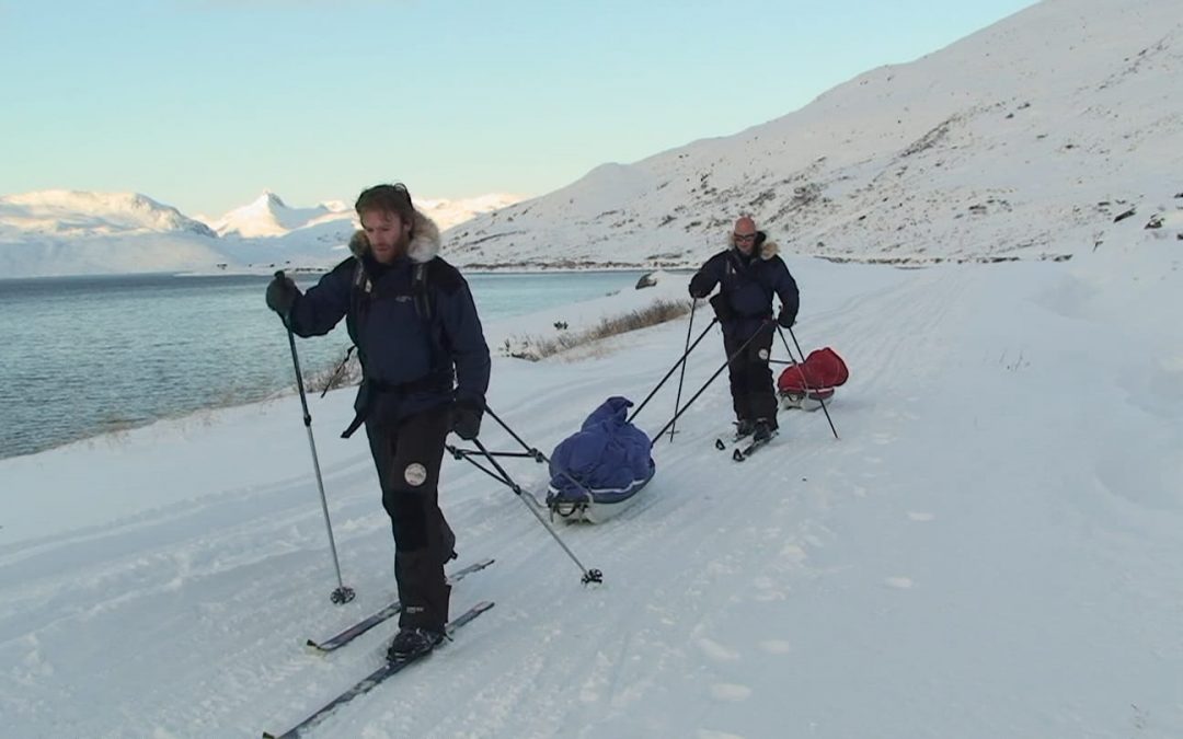 Simon ODonnell stands in front of Mark Pollock as they both ski along the snow pulling their pulks. (Mark is attached to Simons pulk.) Both are dressed in navy and black snow gear with fur hoods on their jackets. Mark wears dark glasses. 