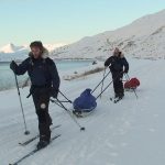 Simon ODonnell stands in front of Mark Pollock as they both ski along the snow pulling their pulks. (Mark is attached to Simons pulk.) Both are dressed in navy and black snow gear with fur hoods on their jackets. Mark wears dark glasses. 