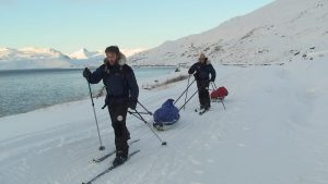 Simon ODonnell stands in front of Mark Pollock as they both ski along the snow pulling their pulks. (Mark is attached to Simons pulk.) Both are dressed in navy and black snow gear with fur hoods on their jackets. Mark wears dark glasses. 