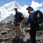 John ORegan and Mark Pollock hike up the side of a mountain as part of the Everest Marathon. John ORegan is to the left of the picture, wearing dark glasses, a white hat and blue and black shirt and shorts. Mark is to the right of the shot and wearing a navy hat, a blue and grey jacket and is carrying a black backpack. John and Mark are both holding onto either end of two poles as they climb.