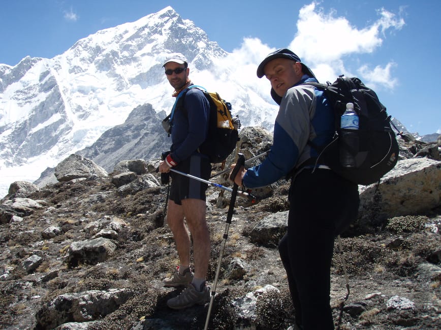 John ORegan and Mark Pollock hike up the side of a mountain as part of the Everest Marathon. John ORegan is to the left of the picture, wearing dark glasses, a white hat and blue and black shirt and shorts. Mark is to the right of the shot and wearing a navy hat, a blue and grey jacket and is carrying a black backpack. John and Mark are both holding onto either end of two poles as they climb.
