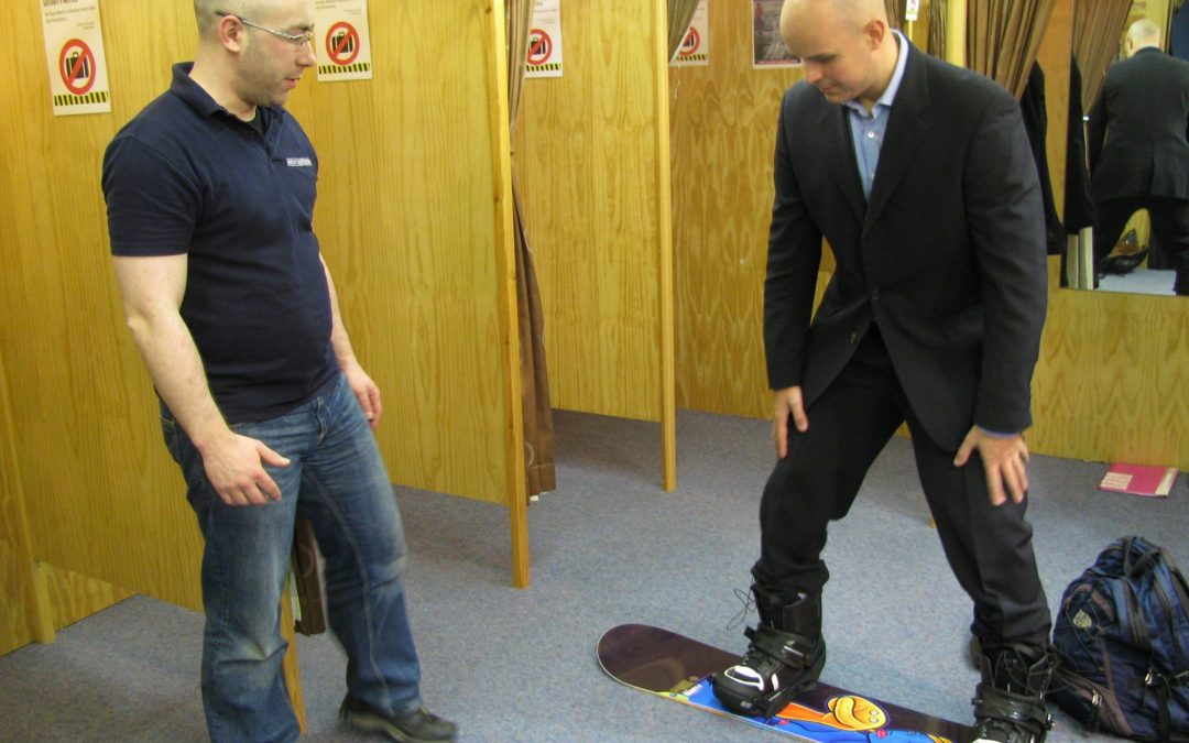 Mark Pollock, dressed in a suit, is standing on a snowboard smiling as he tries it out in a shop. A man to the left looks on. 