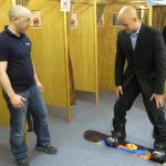 Mark Pollock, dressed in a suit, is standing on a snowboard smiling as he tries it out in a shop. A man to the left looks on. 