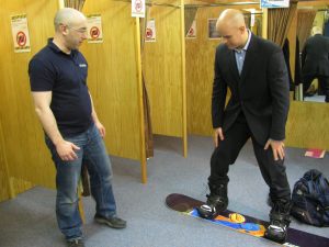 Mark Pollock, dressed in a suit, is standing on a snowboard smiling as he tries it out in a shop. A man to the left looks on. 