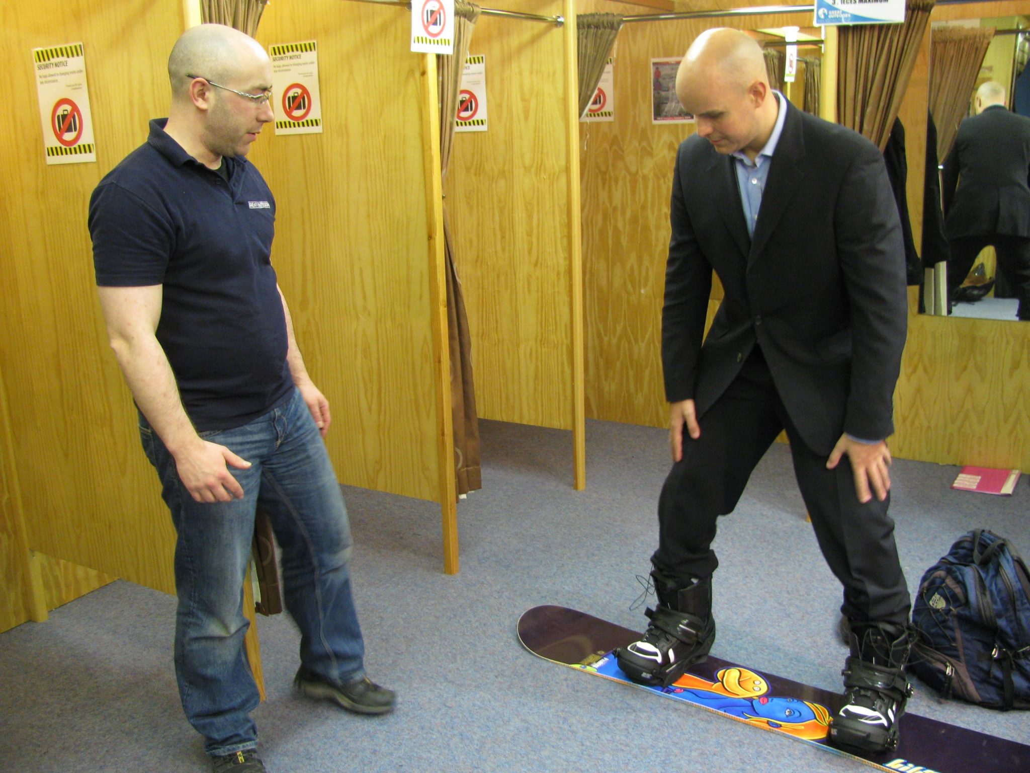 Mark Pollock, dressed in a suit, is standing on a snowboard smiling as he tries it out in a shop. A man to the left looks on. 