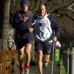 Mark Pollock is running on a quiet footpath with a guide to the left. He wears a grey and navy hoodie and carries his guide stick.