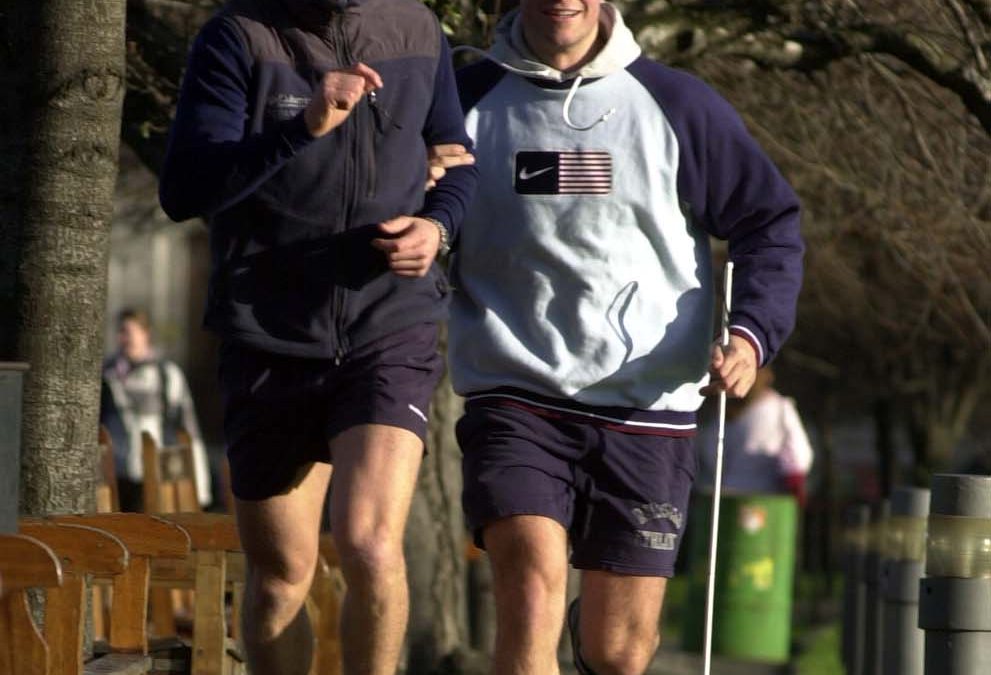 Mark Pollock is running on a quiet footpath with a guide to the left. He wears a grey and navy hoodie and carries his guide stick.
