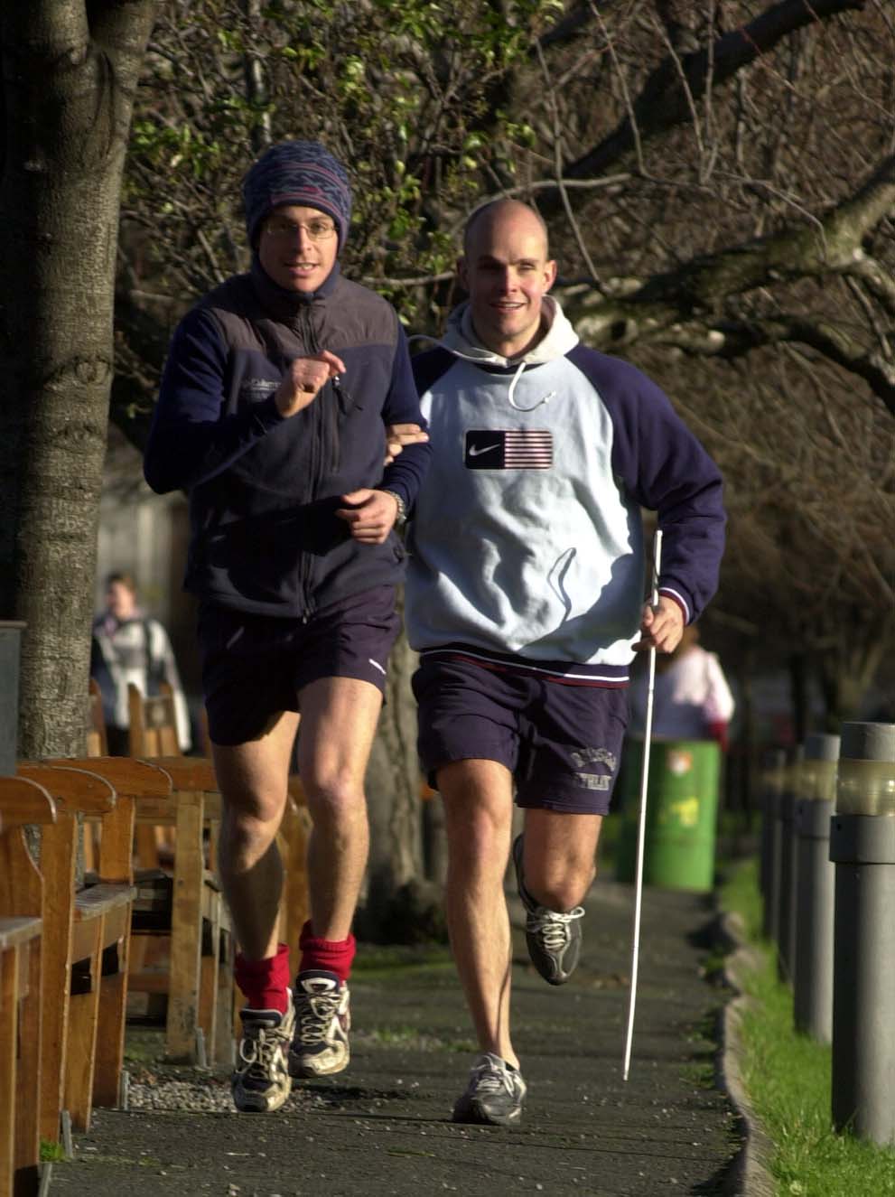 Mark Pollock is running on a quiet footpath with a guide to the left. He wears a grey and navy hoodie and carries his guide stick.