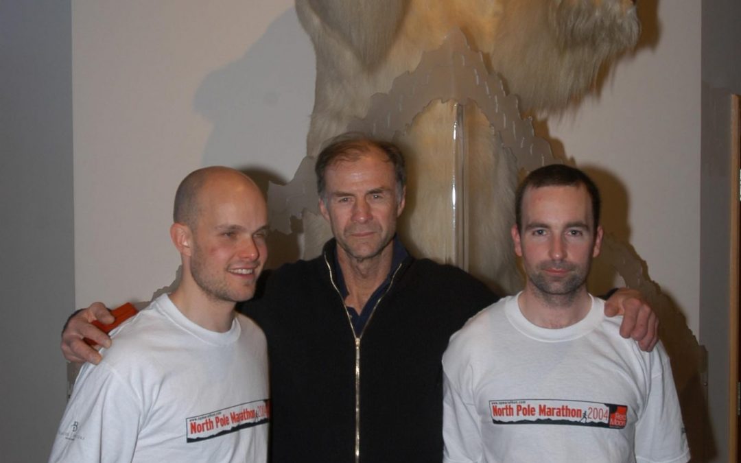 Mark Pollock, Sir Ranulph Fiennes and John ORegan stand in front of a stuffed polar bear. Mark and John both wear a white North Pole Marathon 2004 t-shirt. Ralph stands in between them with his hands on both of their shoulders, wearing a black zip up.