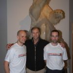 Mark Pollock, Sir Ranulph Fiennes and John ORegan stand in front of a stuffed polar bear. Mark and John both wear a white North Pole Marathon 2004 t-shirt. Ralph stands in between them with his hands on both of their shoulders, wearing a black zip up.