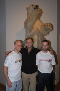 Mark Pollock, Sir Ranulph Fiennes and John ORegan stand in front of a stuffed polar bear. Mark and John both wear a white North Pole Marathon 2004 t-shirt. Ralph stands in between them with his hands on both of their shoulders, wearing a black zip up.