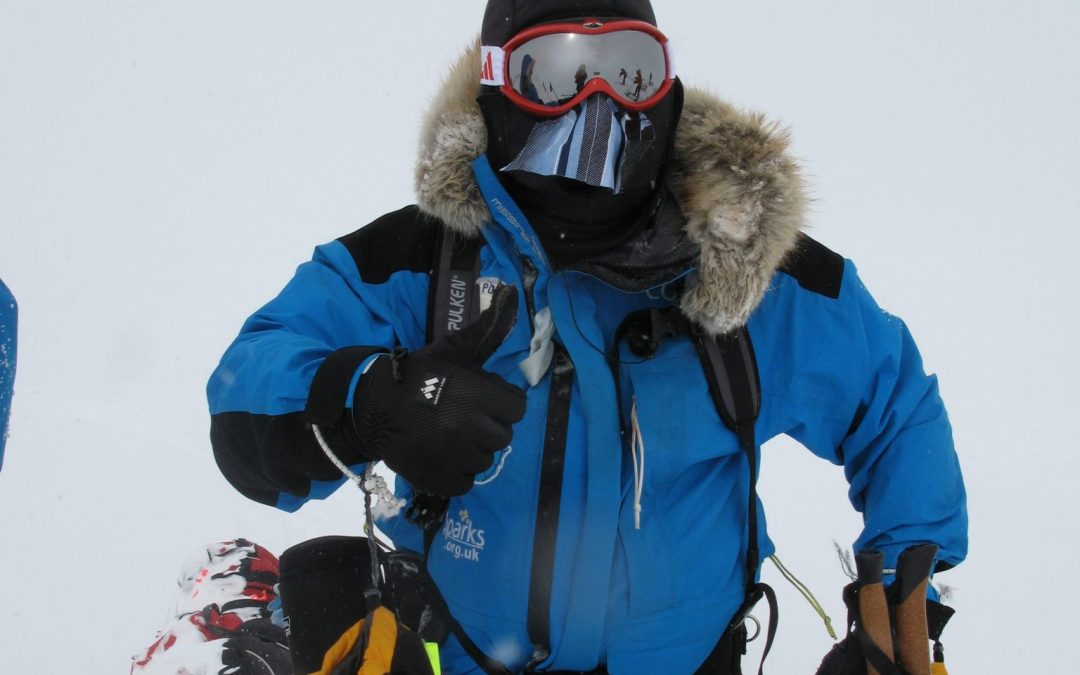 Mid-range shot of Mark Pollock standing in the snow in full snow gear, wearing balaclava, goggles, snow jacket with fur hood. One hand is in a thick yellow gloves while the other glove is removed so he can give a thumbs up gesture.