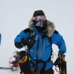 Mid-range shot of Mark Pollock standing in the snow in full snow gear, wearing balaclava, goggles, snow jacket with fur hood. One hand is in a thick yellow gloves while the other glove is removed so he can give a thumbs up gesture.