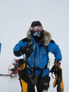 Mid-range shot of Mark Pollock standing in the snow in full snow gear, wearing balaclava, goggles, snow jacket with fur hood. One hand is in a thick yellow gloves while the other glove is removed so he can give a thumbs up gesture.
