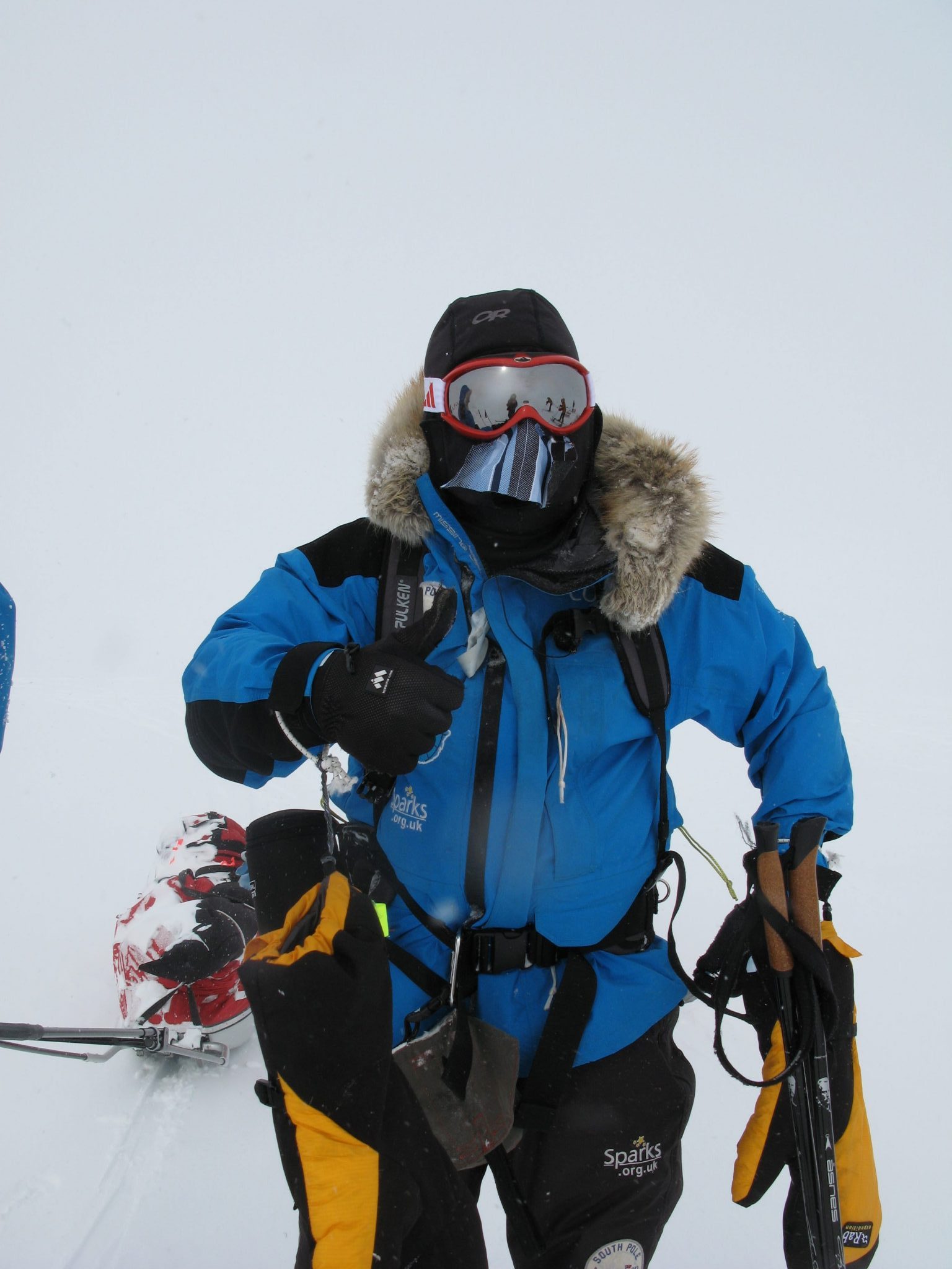 Mid-range shot of Mark Pollock standing in the snow in full snow gear, wearing balaclava, goggles, snow jacket with fur hood. One hand is in a thick yellow gloves while the other glove is removed so he can give a thumbs up gesture.