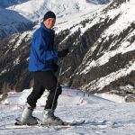 Mark Pollock dressed in a blue snow jacket, black hat, black trousers and silver ski boots as he poses on his skis and smiles at the camera while people ski in the distance behind him.