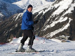 Mark Pollock dressed in a blue snow jacket, black hat, black trousers and silver ski boots as he poses on his skis and smiles at the camera while people ski in the distance behind him.